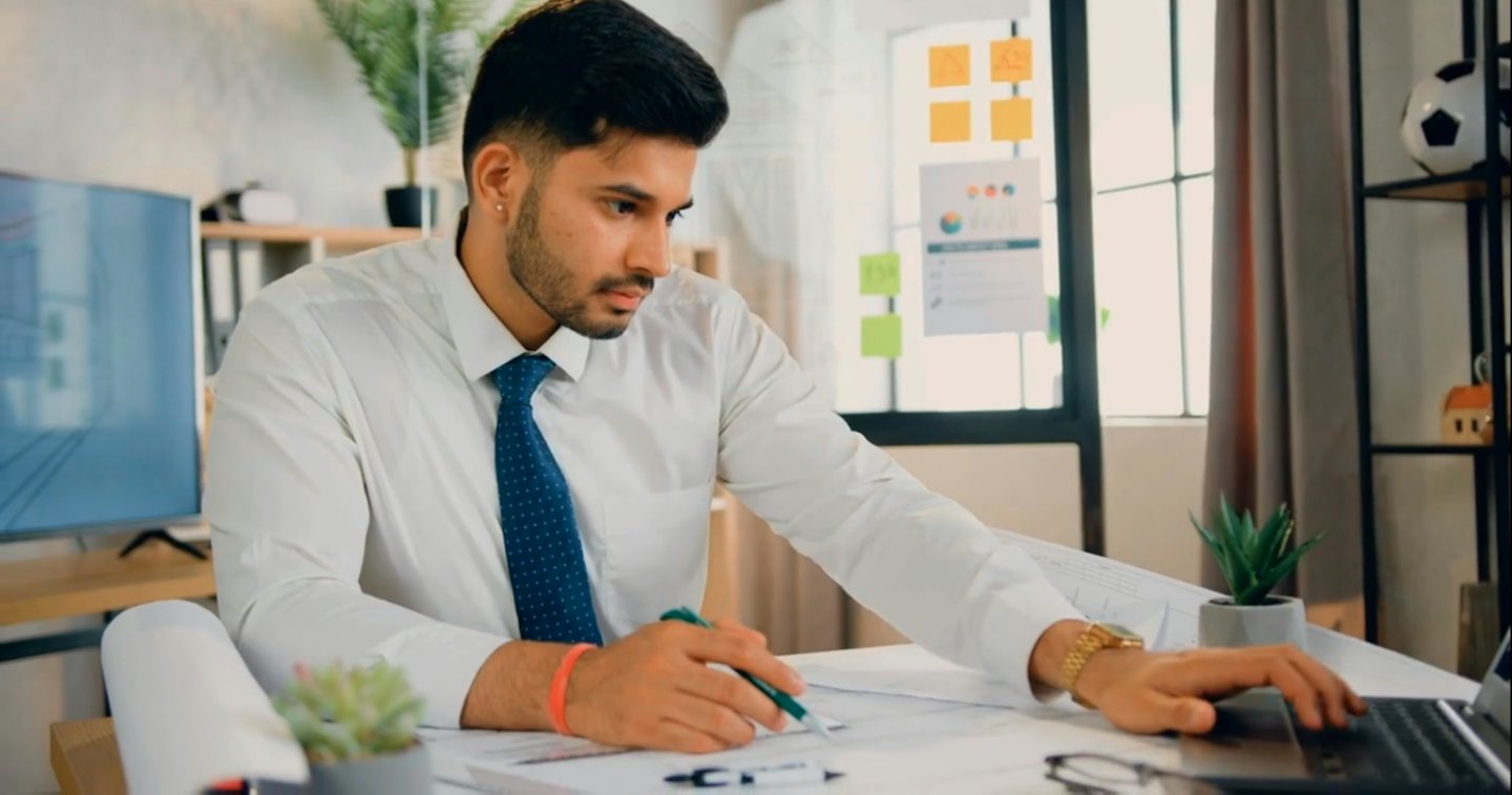 Young man in a white shirt reviews blueprints at a desk.