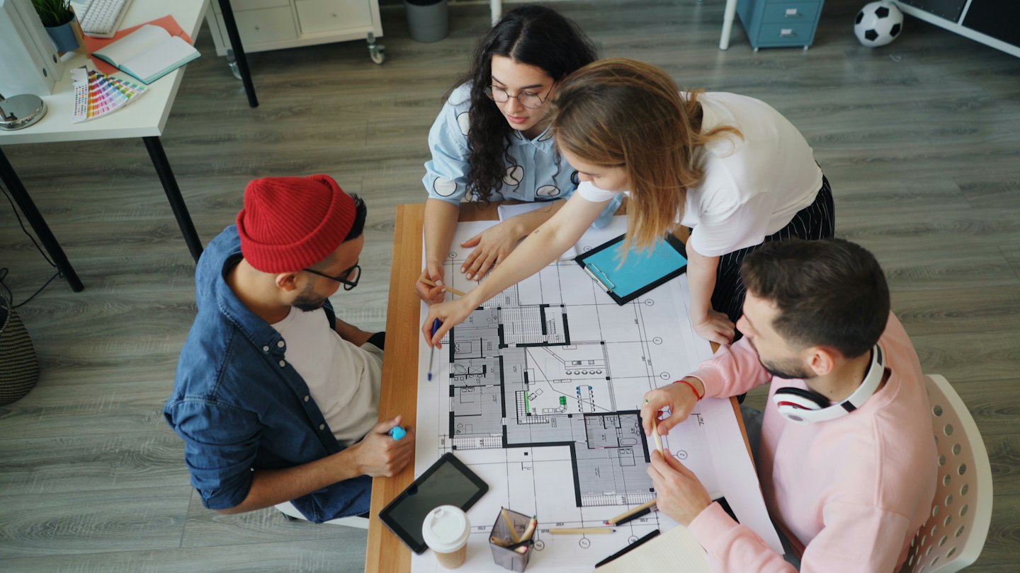 Four designers collaborate over an architectural blueprint on a table.